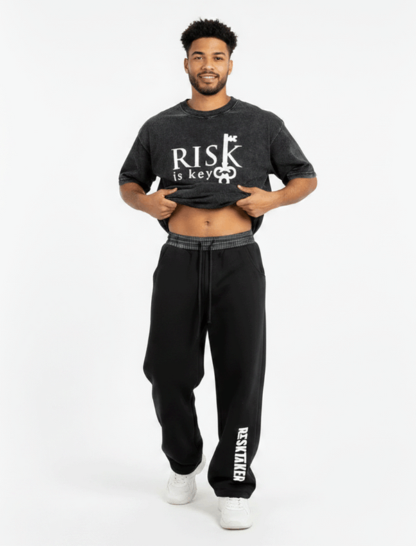 A front-facing studio shot of a model showcasing the unique silhouette of the barrel-leg sweatpants. The model is lifting both sides of their outer top or hands slightly to fully expose the wide, eye-catching black and white plaid contrast waistband. The sweatpants are styled in a loose, oversized fit, showcasing how the heavyweight fleece bows out at the thighs before tapering cleanly at the ribbed ankle cuffs. The model wears the sweatpants pulled up to a mid-rise fit, highlighting the white adjustable dr