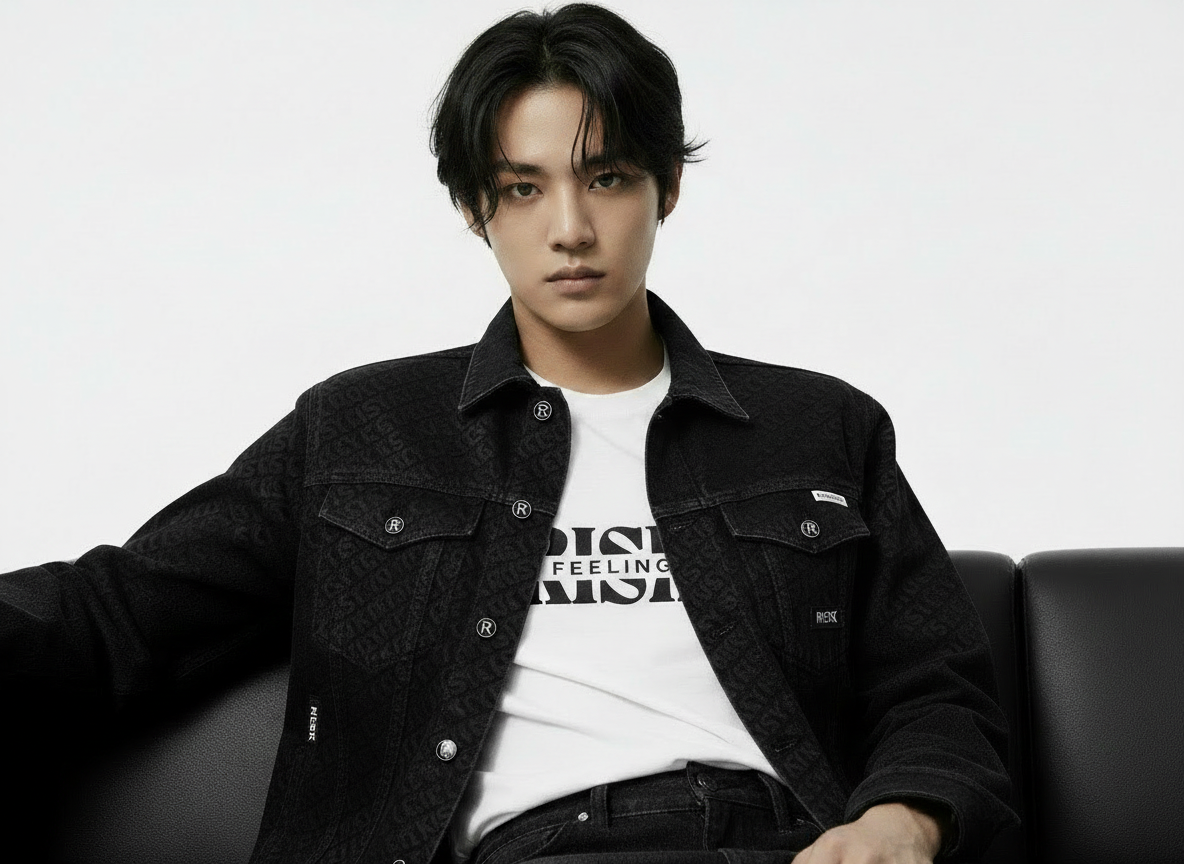 Gen Z male model wearing the black Pack Leader denim logo jacket over a Feeling Riskey t-shirt and black jeans, sitting confidently on a couch.